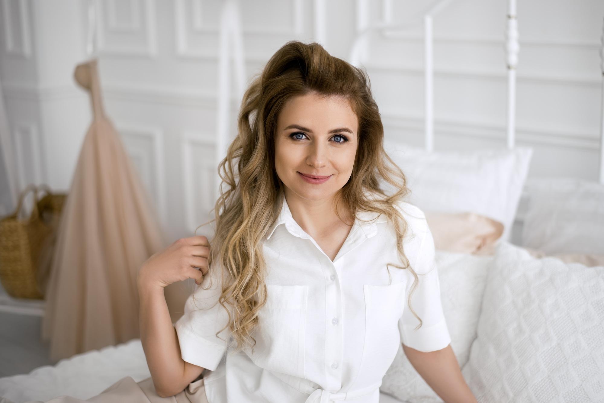 A young, beautiful, curly-haired woman with expressive eyes is sitting on the bed.